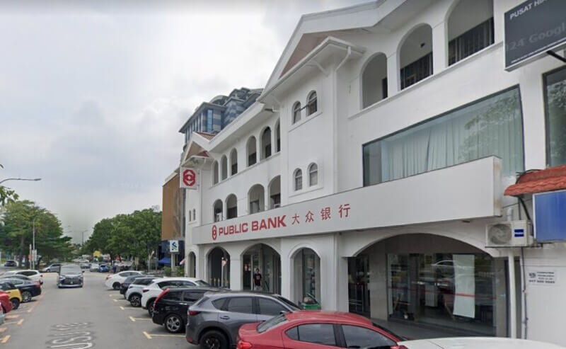 Pandangan luar bangunan cawangan Public Bank UEP Subang Jaya berwarna putih dengan papan tanda dan tulisan Cina, serta deretan kereta yang diletakkan di sepanjang jalan raya.