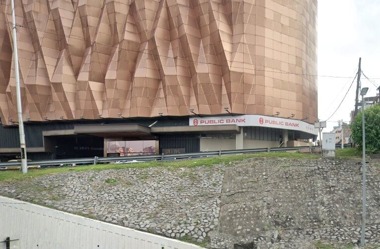 Imej menunjukkan bahagian bawah bangunan besar dengan fasad geometri berwarna gangsa yang tersendiri. Sepanduk mendatar dengan logo dan nama "PUBLIC BANK" kelihatan jelas betul-betul di bawah fasad utama, menunjukkan bahawa sebuah cawangan Public Bank terletak di sana