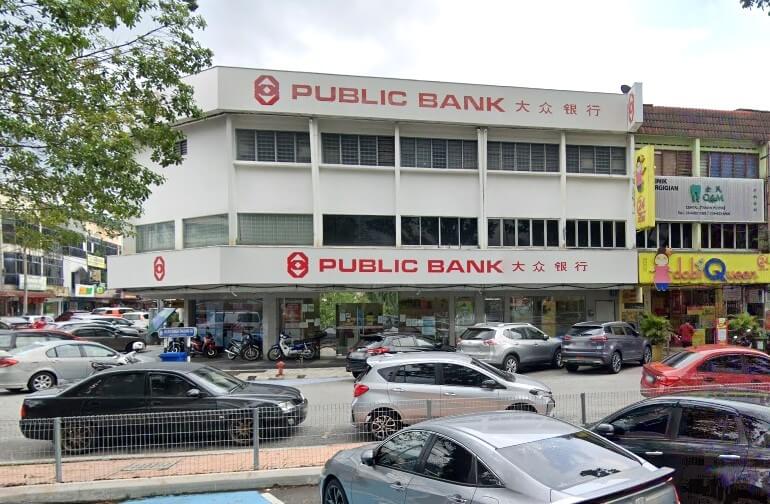 Bahagian luar bangunan cawangan Public Bank Taman Muda, dengan muka bangunan putih dan logo merah, dikelilingi oleh kereta yang diletakkan di kawasan komersial yang sibuk