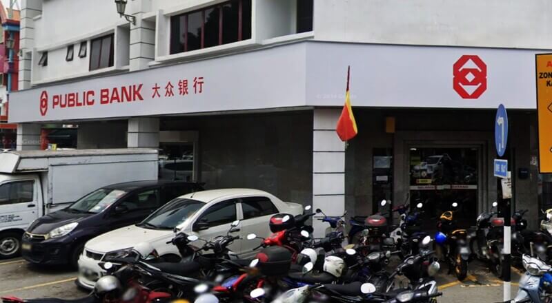 Pandangan hadapan cawangan Public Bank Bandar Sunway dengan papan tanda merah dan cina, bendera Selangor, serta kenderaan dan motosikal yang diletakkan di bahagian hadapan.