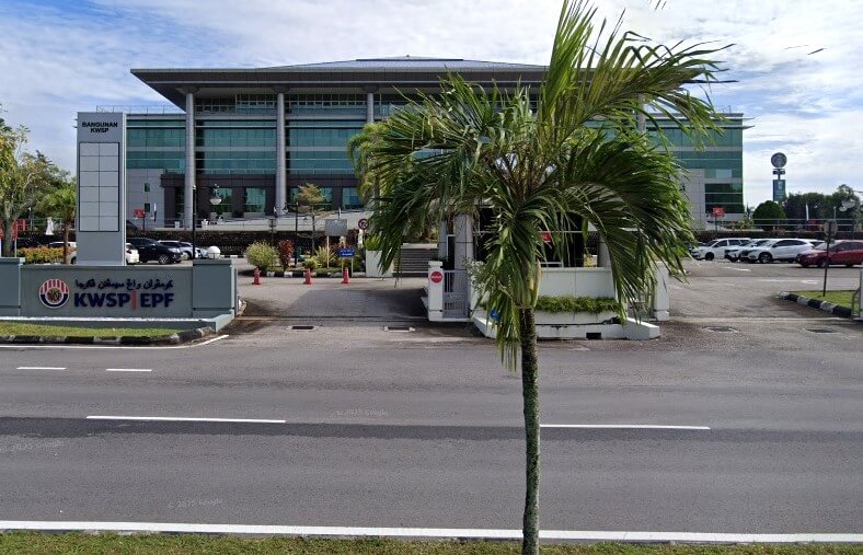 Pemandangan luar yang luas bagi bangunan pejabat Bangunan KWSP (EPF) berdinding kaca moden di Kuantan, Malaysia. Sebatang pokok palem berdiri di latar depan, dan papan tanda besar di sebelah kiri berhampiran pintu masuk bertulis "KWSP EPF" dalam huruf putih dan merah. Bangunan ini dikelilingi oleh kawasan parkir berturap dan jalan raya