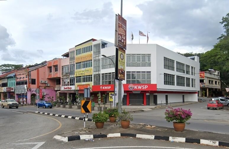 Gambar luaran bangunan pejabat KWSP Raub di Pahang, bangunan moden dua tingkat berwarna putih dengan papan tanda KWSP merah yang mencolok, terletak di sudut jalan bersebelahan dengan rumah kedai lama yang berwarna-warni