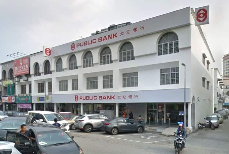 Bahagian luar bangunan cawangan Public Bank Kajang di Selangor, menampilkan fasad putih dengan tingkap melengkung, logo dan nama merah bank dalam bahasa Inggeris dan Mandarin, dan kawasan parkir aras jalan yang sibuk dengan kereta dan motosikal