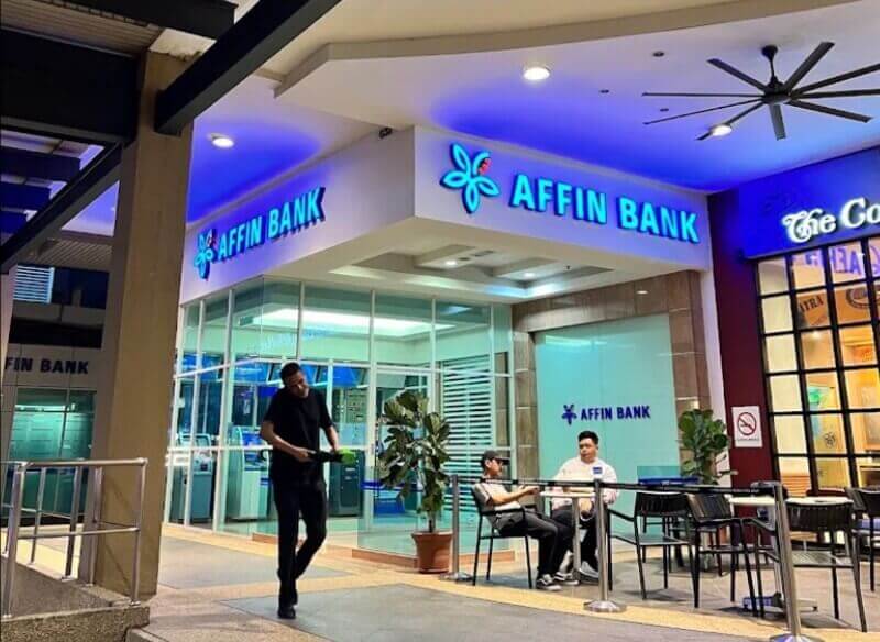 Pandangan hadapan cawangan Affin Bank The Curve pada waktu malam dengan lampu papu tanda biru menyala, serta beberapa orang berdiri dan duduk di kawasan kaki lima berhampiran