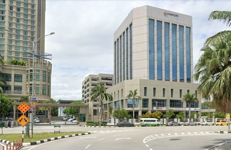 Sebuah bangunan pejabat moden bertingkat berlabel "KWSP EPF" dengan tingkap pemantul biru dan bahagian bawah berwarna krim, terletak bersebelahan dengan persimpangan yang sibuk, dengan pokok palma dan bangunan tinggi lain di sebelah kiri