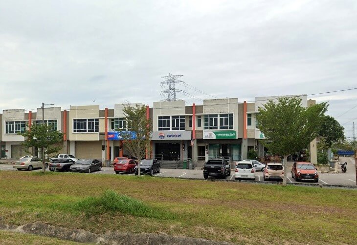 Pandangan luar deretan rumah kedai komersial moden dua tingkat, menunjukkan pintu masuk pejabat KWSP/EPF (Employees Provident Fund) di Alor Gajah, Malaysia, terletak di tengah. Terdapat kereta diletakkan di hadapan kedai dan kawasan berumput di latar depan
