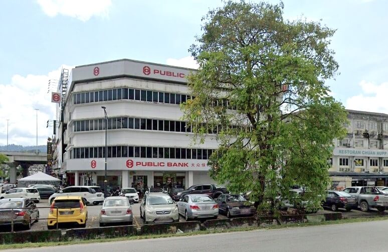 Bahagian luar bangunan cawangan Public Bank Ampang di Selangor, Malaysia dengan kawasan tempat letak kereta yang sibuk dan pokok besar di latar depan