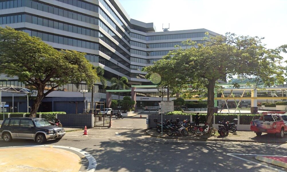 Bangunan pejabat yang besar, moden, bertingkat dengan fasad kaca bertingkat, melengkung, sebahagiannya dikaburkan oleh pokok hijau yang subur di kiri dan kanan. Di latar depan, terdapat kawasan pintu masuk berturap dengan SUV berwarna gelap dan beberapa motosikal parkir. Papan kecil bertulis "KWSP" boleh dilihat berhampiran pintu masuk