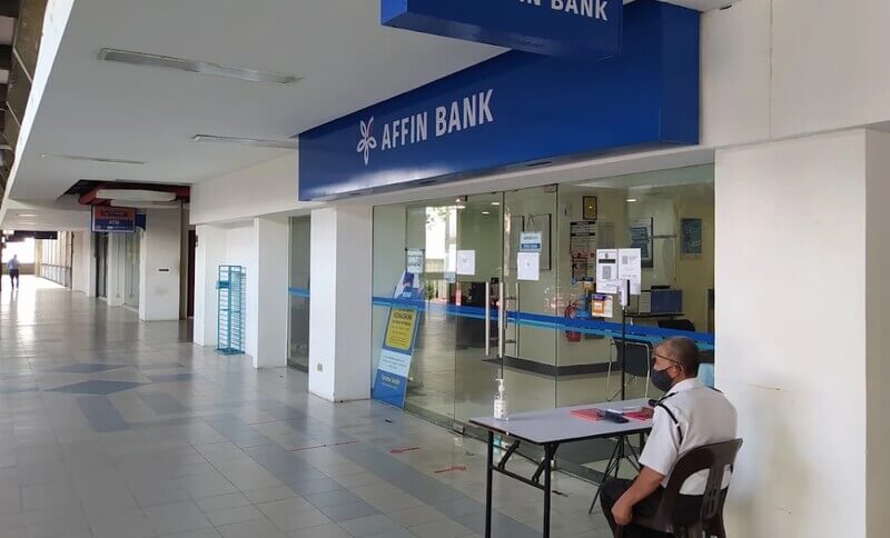 Pintu masuk cawangan Affin Bank di UiTM dengan papan tanda biru bertulis "AFFIN BANK", pintu kaca gelongsor, dan seorang pengawal keselamatan bertopeng muka duduk di meja pendaftaran di hadapan pintu premis.