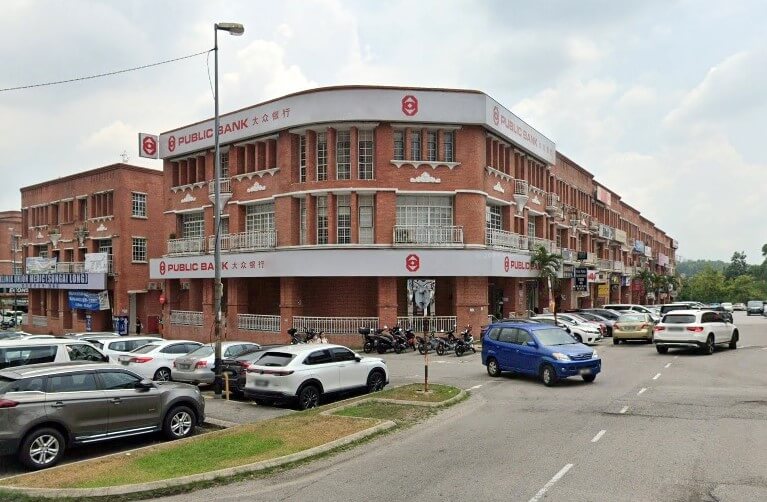 Public Bank Bandar Sungai Long, Selangor, Malaysia. Sebuah bangunan bata besar di sudut jalan dengan kereta diletakkan dan sebuah van biru di latar depan