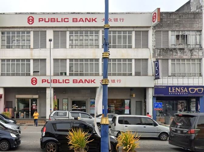 Pemandangan luar cawangan Public Bank di kawasan komersial, dengan kereta diletakkan di latar depan dan kedai Lensa Optometry di sebelahnya