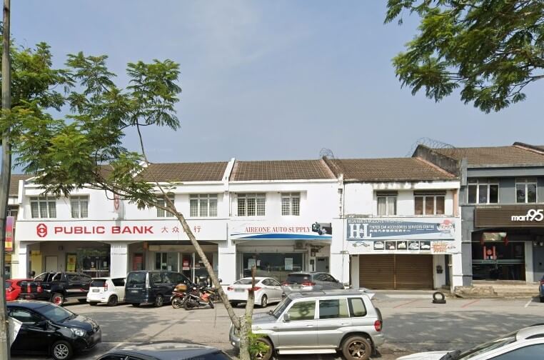 Pemandangan jalanan deretan rumah kedai komersial putih dua tingkat, dengan cawangan Public Bank menduduki unit sebelah kiri, ditandakan dengan logo merah dan nama dalam skrip Inggeris dan Mandarin. Kedai-kedai lain, seperti kedai bekalan automotif, kelihatan di sebelahnya, dengan kenderaan diletakkan di hadapannya