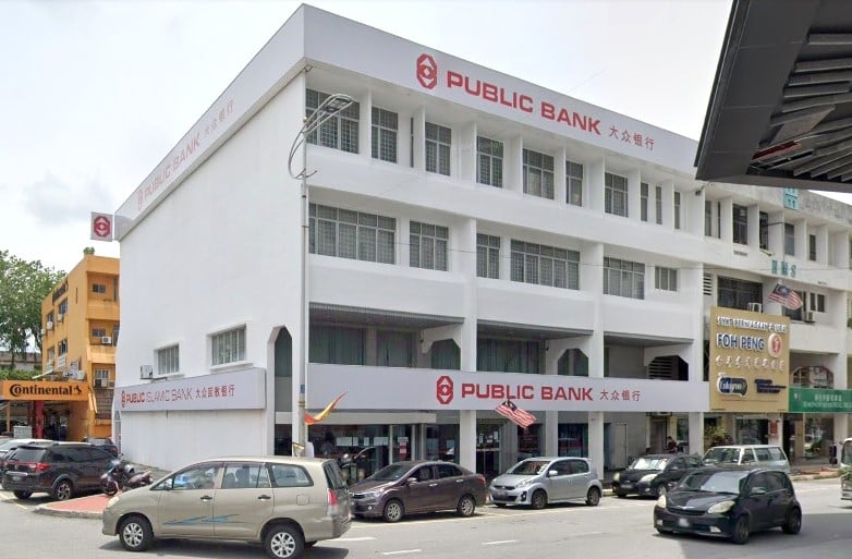 Sebuah bangunan komersial empat tingkat putih yang besar dengan papan tanda Public Bank merah yang mencolok di sepanjang tingkat atas dan bawah. Bangunan ini terletak di sudut jalan dengan lalu lintas dan kereta yang diletakkan di latar depan, bersebelahan dengan rumah kedai lain seperti Foh Peng