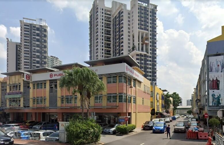 Bangunan cawangan Public Bank tiga tingkat, dengan panel fasad merah jambu dan kuning, dengan jalan yang sibuk dan kawasan tempat letak kereta di latar depan. Beberapa menara kondominium moden berdiri di latar belakang