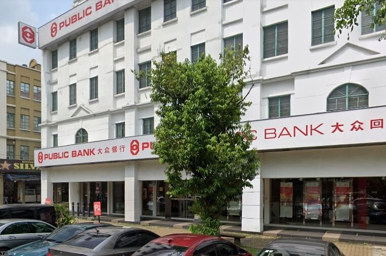 Bahagian luar bangunan Public Bank Kota Kemuning, bangunan bertingkat berwarna putih dengan papan tanda merah, sebahagiannya dikaburkan oleh pokok hijau di tengah, dengan kereta diletakkan di latar depan