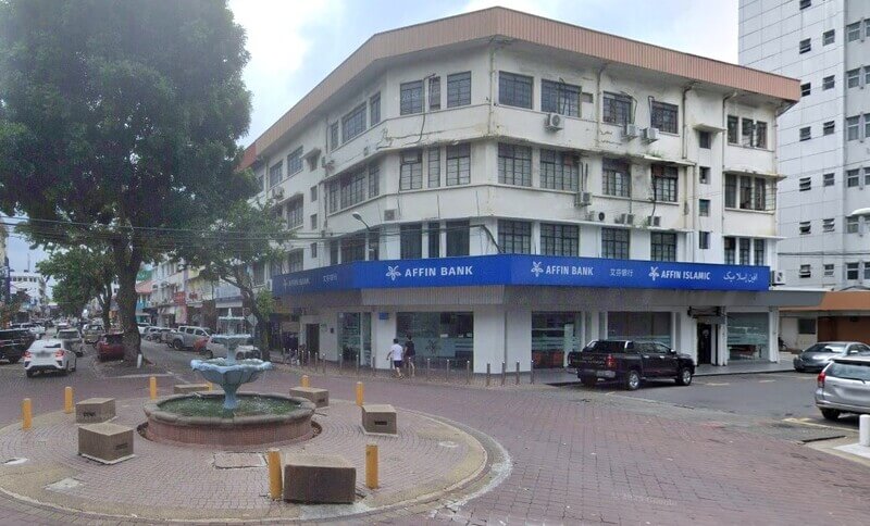 Pemandangan luar cawangan Affin Bank bertingkat berwarna putih yang terletak di sudut jalan bersebelahan dengan air pancut batu kecil dan laluan pejalan kaki berturap