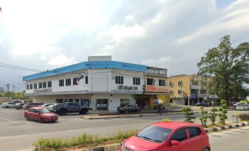 Pandangan luar bangunan RHB Bank dua tingkat berwarna putih di Kuala Dungun, memaparkan papan tanda biru di sudut jalan dengan kereta yang diletakkan