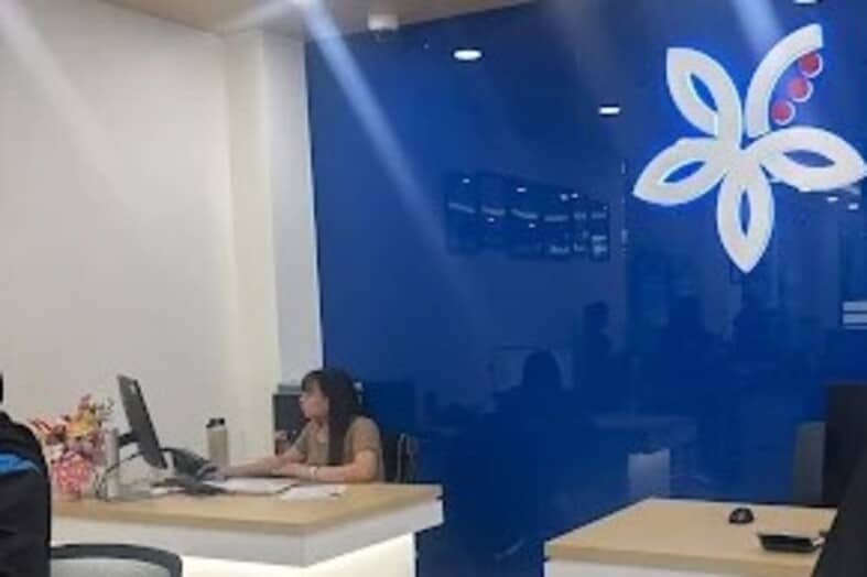 Pandangan dalaman cawangan AFFIN Bank, menunjukkan seorang pekerja wanita duduk di meja kayu ringan, bekerja pada komputer. Latar belakangnya memaparkan dinding biru yang menyerlah dengan logo AFFIN Bank berwarna putih dan merah besar dengan lima kelopak