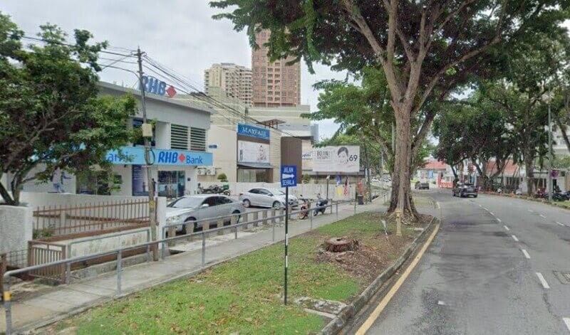 Pemandangan jalan raya di hadapan cawangan RHB Bank Jalan Burma dengan papan tanda jalan, pokok-pokok hijau, dan beberapa kenderaan di tepi jalan