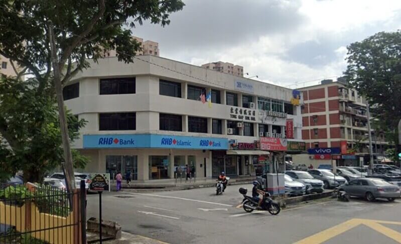 Pandangan luar bangunan sudut tiga tingkat di Jelutong yang menampilkan Bank RHB dan cawangan RHB Islamic di tingkat bawah, dengan tempat letak kereta tepi jalan dan penunggang motosikal yang lalu-lalang