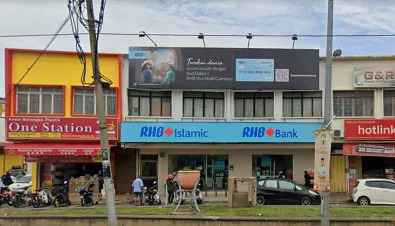Pandangan luar cawangan RHB Bank Rantau dan RHB Islamic yang terletak di deretan lot kedai. Papan iklan hitam besar untuk Kad Debit Pelbagai Mata Wang RHB Visa dipasang di atas papan tanda hadapan kedai berwarna biru. Kedai-kedai bersebelahan termasuk "One Station" di sebelah kiri dan pengedar "Hotlink" di sebelah kanan