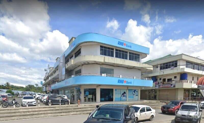 Bangunan RHB Bank tiga tingkat di Keningau dengan papan tanda biru muda dan sudut bulat, menampilkan pemandangan aras jalan dengan kereta yang diletakkan