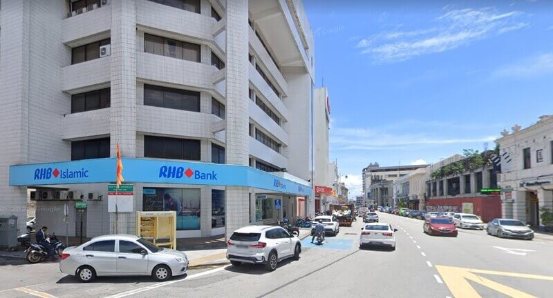 Pandangan luar sebuah bank RHB bertingkat dan cawangan RHB Islamic yang terletak di sudut jalan di Lebuh Pantai, Pulau Pinang, menampilkan seni bina jubin putih di bawah langit biru yang cerah dengan kereta-kereta yang diletakkan dan dipandu di sepanjang jalan