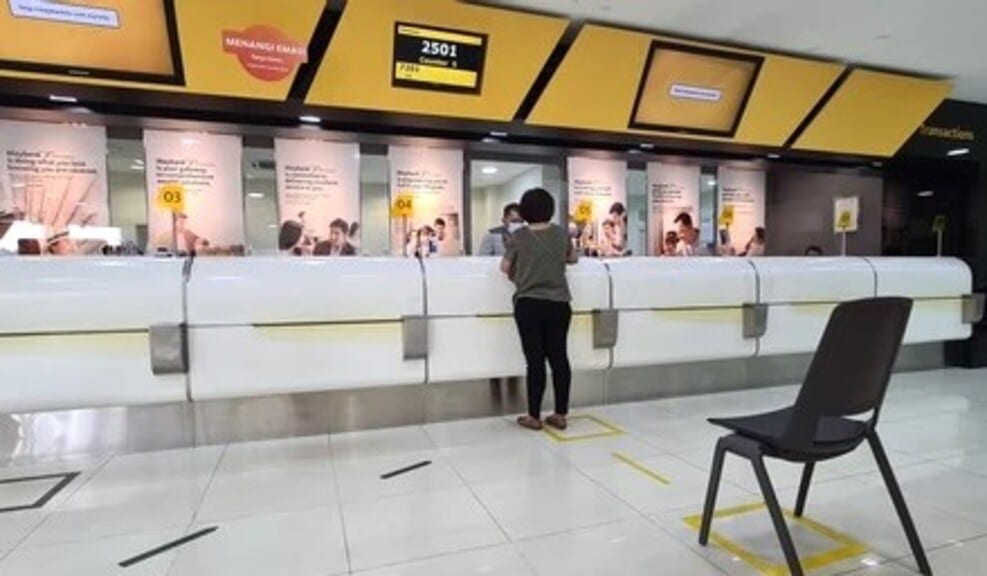 Seorang wanita berdiri di kaunter perkhidmatan Maybank moden di Kuala Lumpur, menampilkan penjenamaan kuning dan putih, paparan baris gilir digital menunjukkan nombor 2501, dan penanda lantai jarak sosial