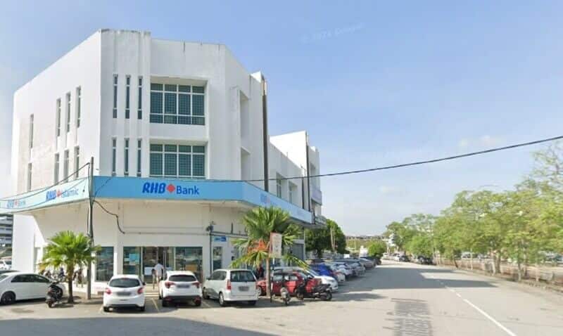 Pandangan luar cawangan RHB Bank tiga tingkat berwarna putih yang terletak di lot sudut di Bukit Mertajam, memaparkan papan tanda "RHB Bank" berwarna biru muda dan kereta-kereta yang diletakkan di sepanjang jalan