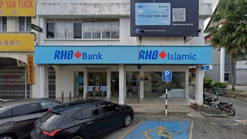 Papan tanda berwarna biru cerah memaparkan logo "RHB Bank" dan "RHB Islamic" di bahagian hadapan cawangan bank pada bangunan dua tingkat. Di atasnya terdapat papan iklan papan tanda kad debit RHB