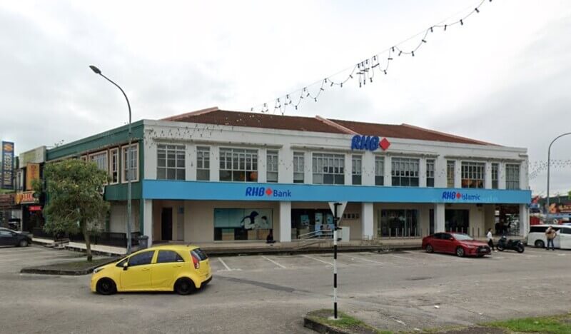 Pandangan luar bangunan dua tingkat RHB Bank dan cawangan RHB Islamic di Bandar Baru Uda, menampilkan dinding putih, papan tanda biru dan kawasan parkir dengan kereta kuning di latar depan