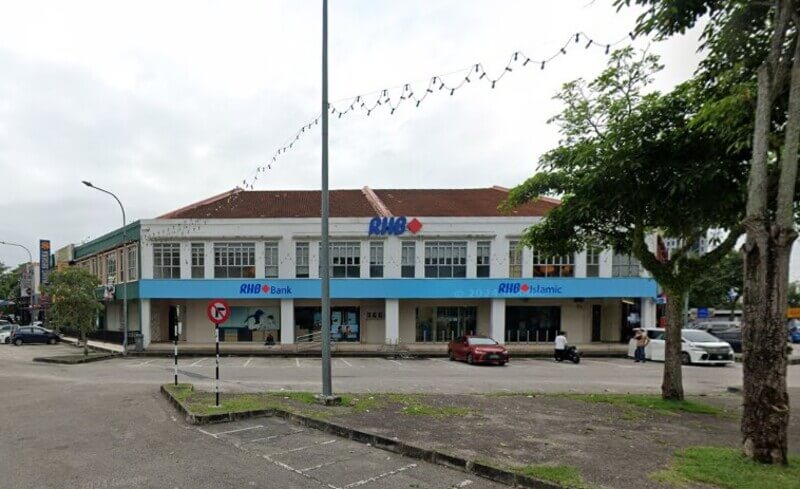 Pemandangan hadapan bangunan cawangan RHB Bank Bandar Baru Uda dengan papan tanda biru yang jelas, berhadapan dengan kawasan tempat letak kereta