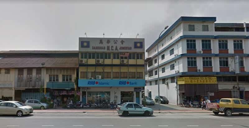 Pemandangan aras jalan Bank RHB dan cawangan RHB Islamic yang terletak di tingkat bawah bangunan Bangunan M.C.A. Jementah berwarna kuning, diapit oleh rumah kedai tradisional dan bangunan putih empat tingkat moden di sebuah pekan di Malaysia