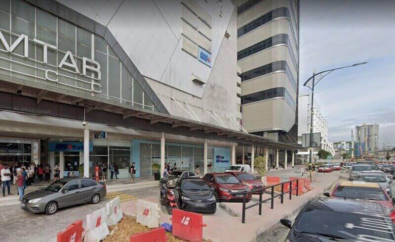 Pemandangan luar cawangan RHB Bank yang terletak di Komtar JBCC di Johor Bahru, menampilkan papan tanda biru bank, pintu masuk aras jalan dengan pejalan kaki, dan jalan yang sibuk dengan kereta yang diletakkan di latar depan