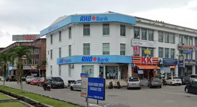 Pemandangan luar bangunan RHB Bank di Kota Tinggi tiga tingkat di sudut jalan dengan kedai KK Super Mart di sebelahnya dan kenderaan di jalan raya