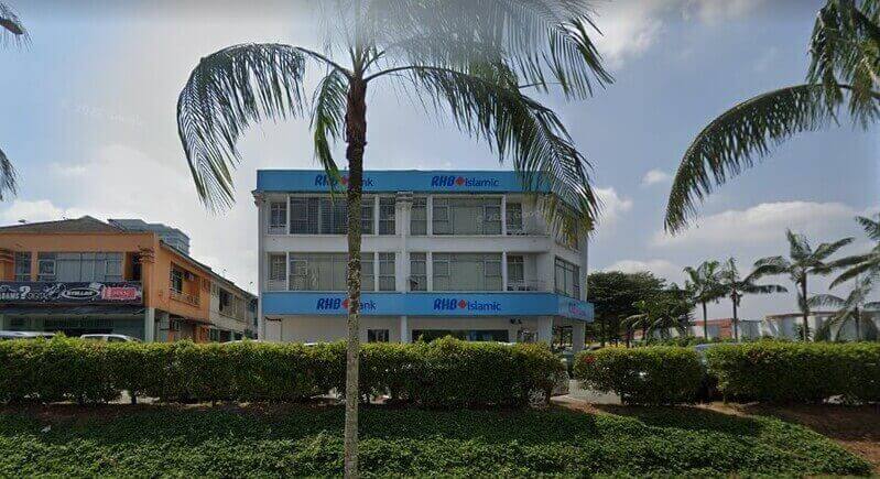 Bangunan tiga tingkat Bank RHB dan cawangan RHB Islamic dengan papan tanda biru, dilihat dari seberang jalan melalui pokok palma dan pagar hijau di bawah langit yang cerah