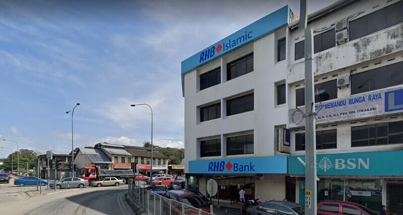 Pandangan luar bangunan sudut bertingkat putih di Ulu Tiram yang menampilkan papan tanda RHB Bank dan RHB Islamic, terletak di sebelah cawangan BSN di jalan yang sibuk dengan lalu lintas