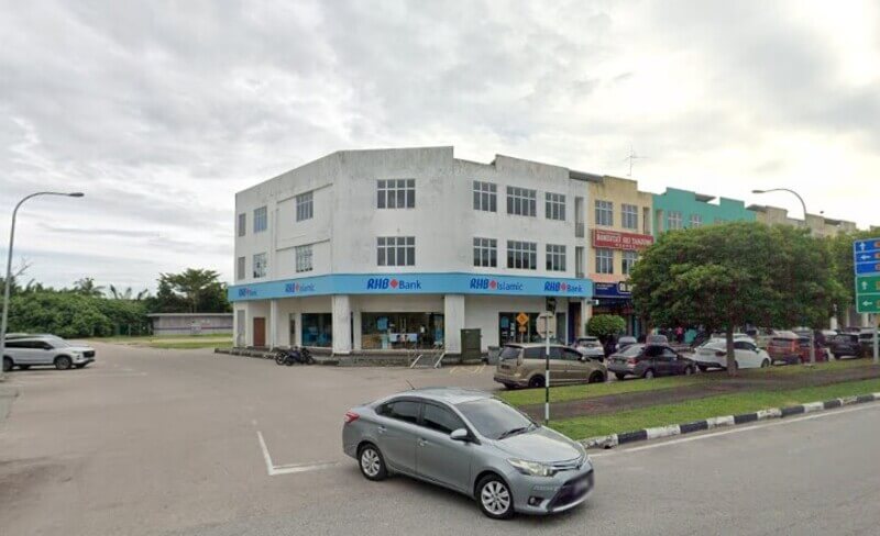 Pandangan sudut bangunan kedai tiga tingkat yang menempatkan cawangan RHB Bank dan RHB Islamic dengan papan tanda biru. Terdapat beberapa buah kereta diletakkan di tempat letak kereta di hadapan bangunan dan sebuah kereta perak memandu di jalan raya utama