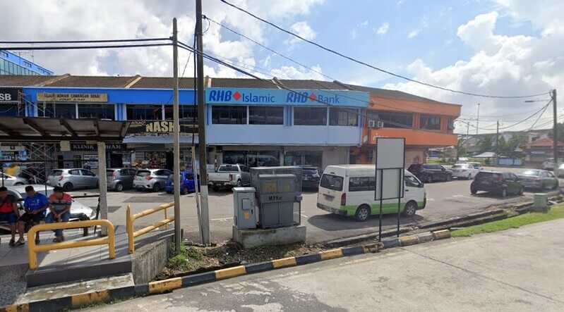 Pemandangan kawasan deretan kedai dua tingkat yang menempatkan cawangan RHB Bank Senai pada hari yang cerah. Kelihatan beberapa orang sedang duduk santai di perhentian bas di sebelah kiri, dan banyak kenderaan diparkir di kawasan hadapan kedai