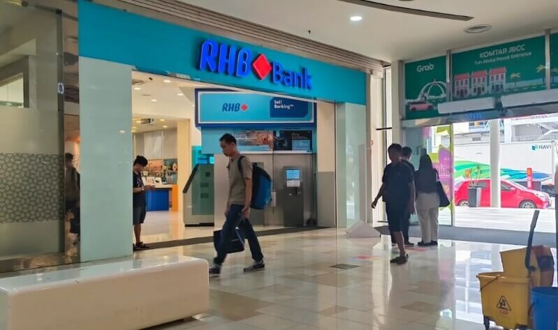 Pintu masuk cawangan RHB Bank Komtar JBCC berlatar belakangkan papan tanda berwarna biru terang di dalam kompleks KOMTAR JBCC. Kelihatan kawasan Perbankan Layan Diri (Self-Banking) di dalam kedai dan beberapa pengunjung sedang berjalan di ruang pejalan kaki.