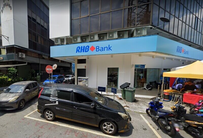 Pandangan luar cawangan RHB Bank Ampang Pointdengan papan tanda biru, kenderaan yang diletakkan di hadapan bangunan, dan khemah perniagaan di sebelah kanan