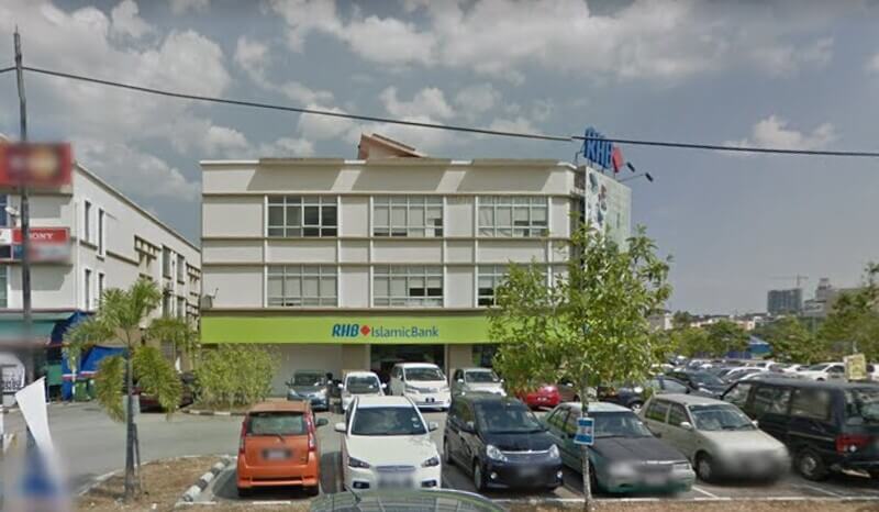 Pandangan hadapan cawangan cawangan RHB Islamic Bank di bangunan kedai tiga tingkat, dengan papan tanda hijau bertulis "RHB Islamic Bank" di aras bawah. Di bahagian hadapan bangunan terdapat kawasan tempat letak kereta yang dipenuhi dengan beberapa buah kenderaan dan pokok-pokok renek.