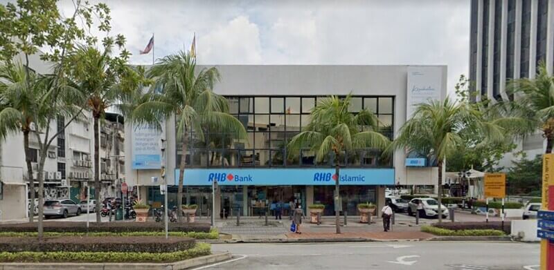 Bangunan cawangan RHB Bank dan RHB Islamic dua tingkat dengan papan tanda biru di hadapan, dikelilingi pokok kelapa sawit/hiasan di tepi jalan raya
