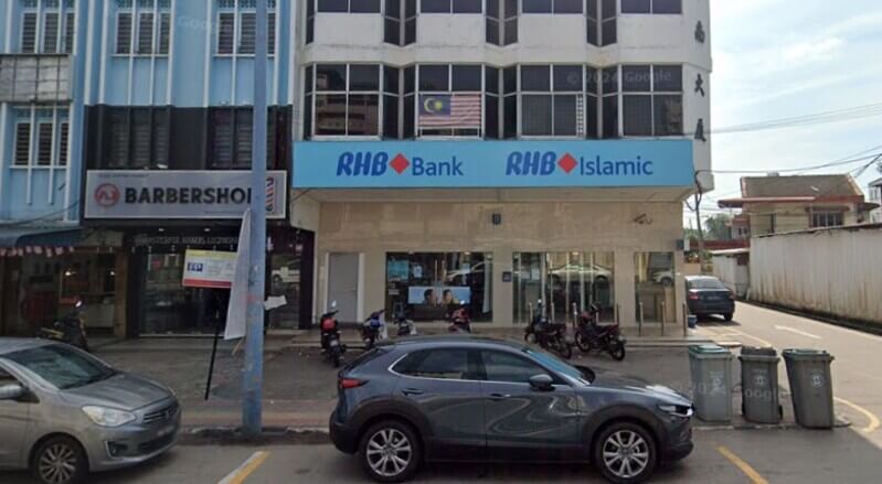 Pandangan hadapan cawangan RHB Bank Tangkak dan RHB Islamic di cawangan kedai, lengkap dengan papan tanda biru, bendera Malaysia di tingkat atas, serta kenderaan yang diletakkan di bahagian hadapan.