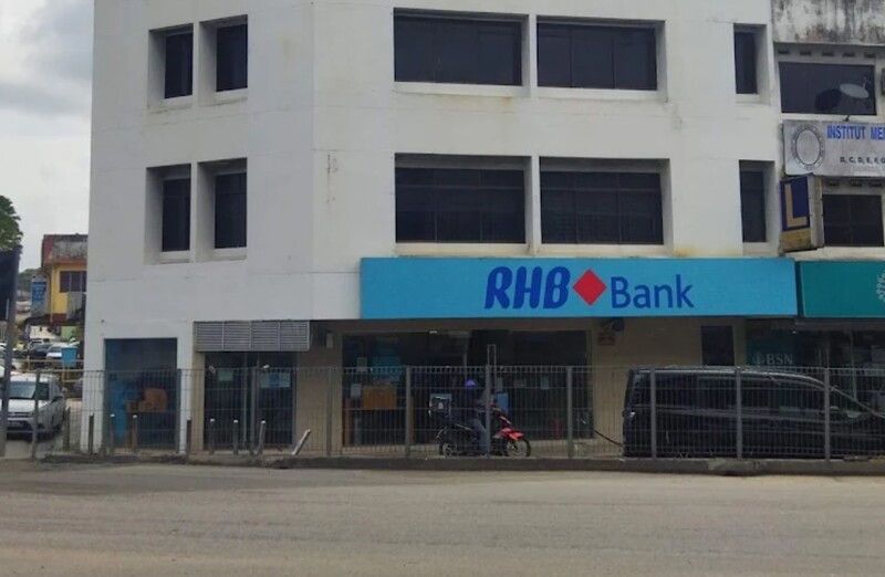 Bangunan putih beberapa tingkat yang menempatkan cawangan RHB Bank di tingkat bawah dengan papan tanda biru bertulis "RHB Bank". Terdapat pagar besi, jalan raya di hadapan, serta sebuah motosikal dan kenderaan yang diletakkan berhampiran