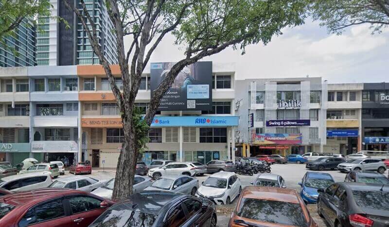 Pemandangan luaran deretan bangunan komersial pada waktu siang yang menempatkan cawangan RHB Bank dan RHB Islamic. Terdapat sebuah papan iklan besar di atas bangunan bank tersebut, sebatang pokok besar di bahagian hadapan, serta kawasan tempat letak kereta awam yang penuh dengan pelbagai jenis kenderaan.