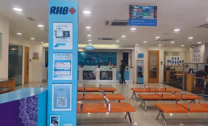 Suasana dalaman cawangan RHB Bank Setia Alam dengan tiang paparan maklumat berwarna biru, kerusi menunggu berwarna jingga, dan kaunter perkhidmatan pelanggan