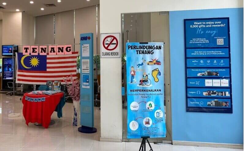 Bahagian dalam cawangan RHB Bank First Avenue memaparkan paparan insurans "Perlindungan Tenang", bendera Malaysia yang dipasang di atas meja dan poster program ganjaran biru di dinding