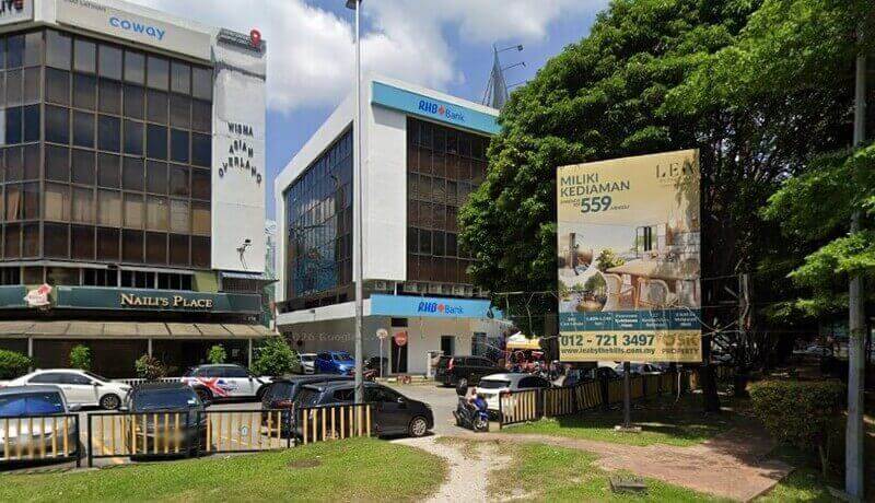 Pemandangan aras jalan cawangan RHB Bank yang terletak di bangunan bertingkat putih bersebelahan Wisma Asian Overland dan Naili's Place. Sebuah papan iklan hartanah berwarna kuning yang besar berdiri di latar depan, dan beberapa buah kereta diletakkan di sepanjang jalan