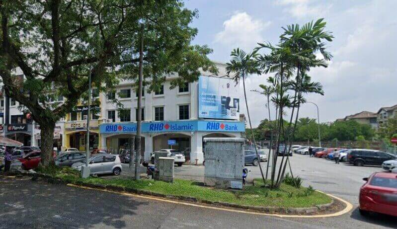 Pemandangan aras jalan Bank RHB dan cawangan RHB Islamic di Bandar Sri Damansara, yang terletak di bangunan sudut tiga tingkat berwarna putih dengan papan tanda biru. Latar depan menunjukkan jalan berturap, tepi jalan berumput dengan pokok palma, dan beberapa kereta yang diletakkan di tempat